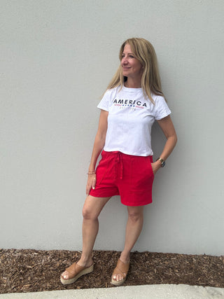 Woman wearing a white t-shirt with 'America' printed on it and red shorts against a gray wall.