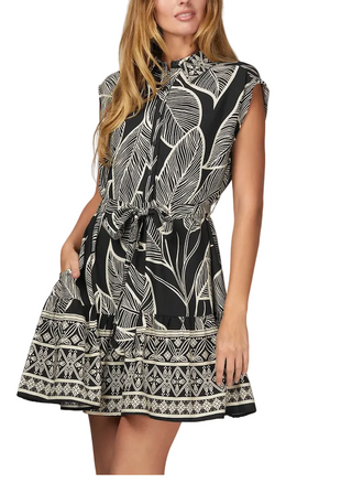 A woman wearing a sleeveless black dress with a white tropical leaf print and a patterned border on the skirt, photographed against a light gray studio background.
