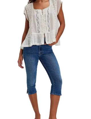 A woman wearing a white sleeveless lace-trim blouse and blue cropped denim jeans, shown from the shoulders down against a light gray studio background.