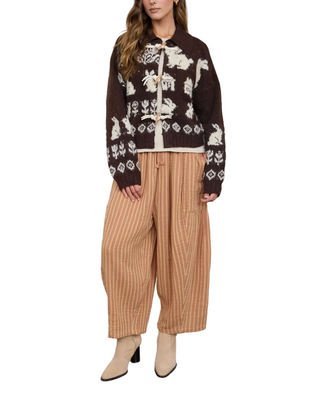 A model wearing a dark brown and cream patterned knit cardigan, beige pinstriped wide-leg trousers, and beige ankle boots, standing against a plain light gray wall.