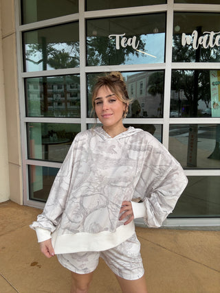Person wearing a white patterned hoodie and shorts standing in front of a store with large windows in a Camo Print Hoodie
