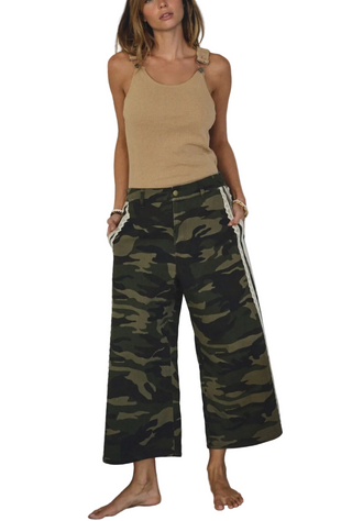 Woman wearing a beige tank top and camouflage pants with white stripes in a minimalistic setting.