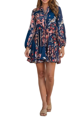 A woman models a navy blue long-sleeve mini dress with a vibrant pink, red, and white floral print, leaning against a textured beige concrete wall.