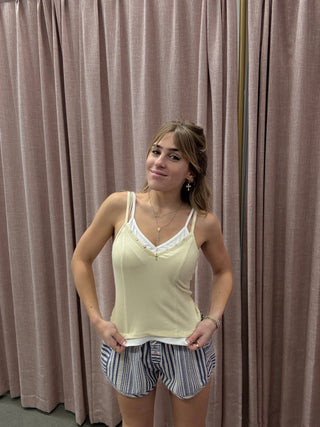 Woman wearing a yellow tank top and striped shorts standing against a pleated curtain.