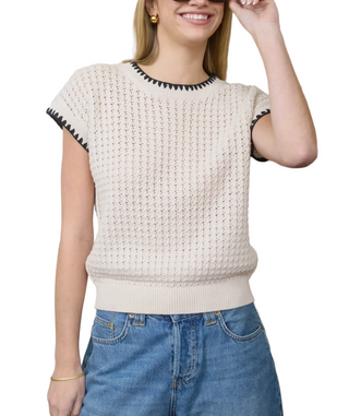 Woman wearing a beige knitted top with black trim and blue jeans against a neutral background