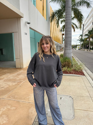 Woman in a gray sweater and blue jeans standing on a sidewalk with palm trees in the background.  Crewneck Ribbed Long Sleeve Knit Top Charcoal