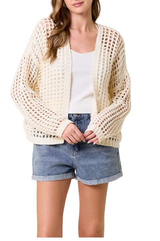 Woman wearing a white crochet cardigan over a white top and denim shorts on a beige background
