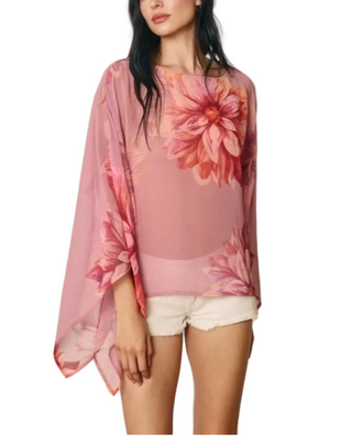 A woman wearing a sheer pink long-sleeve top with a large floral print in shades of pink, red, and peach, paired with white shorts, against a plain light gray background.