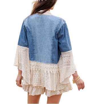 A woman viewed from behind wearing a color-block denim jacket with cream lace ruffle sleeves and hem, standing in a dry grassy field.