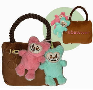 Two plush toys, one pink and one green, hanging from a brown bag with visible branding.