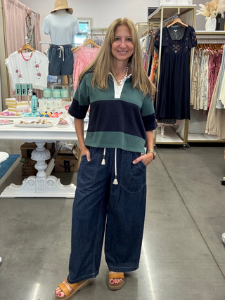 A woman stands in a boutique clothing store wearing a green and navy color-block short-sleeve polo shirt with white drawstrings and dark blue wide-leg denim pants.