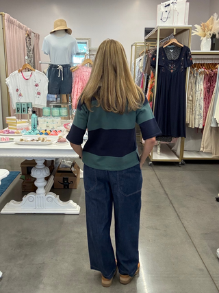 A woman viewed from behind wearing a teal and navy striped short-sleeve top with wide-leg blue jeans, standing inside a boutique clothing store.