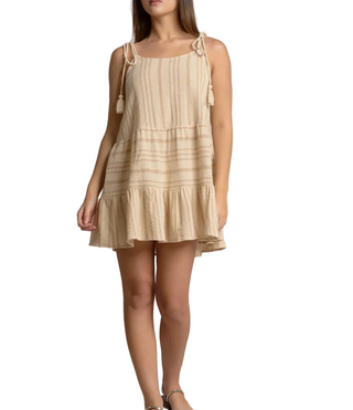 A woman models a sleeveless tiered mini dress in a light beige linen-like fabric with subtle horizontal stripes and tassel shoulder ties, set against a textured dusty pink studio backdrop with a small wooden side table holding two vases.