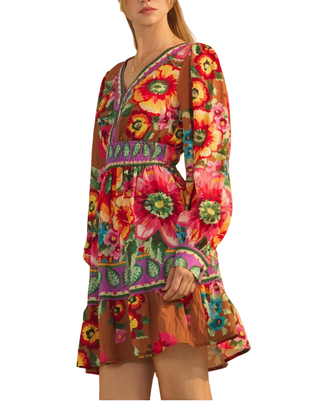 A woman wearing a long-sleeve mini dress with a vibrant multicolor floral print and a brown sheer underlayer, set against a plain beige wall.