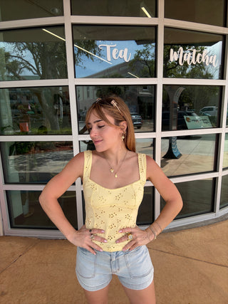 Woman in a yellow top and denim shorts leaning against a glass door with a reflection of trees and a building.