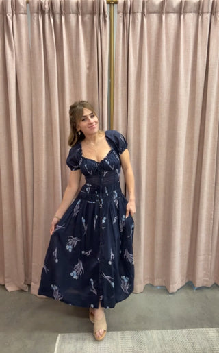 Woman wearing a navy blue dress with white floral patterns against a beige curtain background