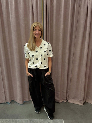 Person wearing a polka dot top and black pants standing in front of a beige curtain.