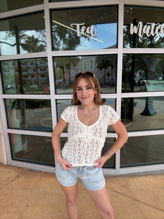 Woman in a white lace top and denim shorts standing in front of a store with large windows.