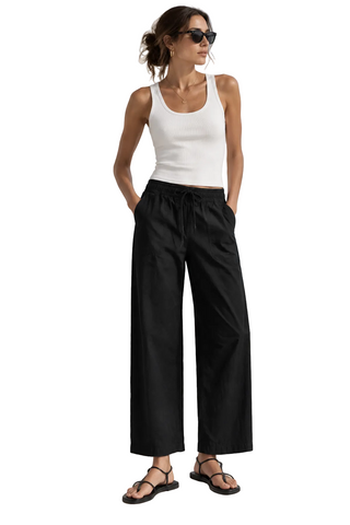A model wearing a beige ribbed tank top and dark navy wide-leg cropped pants with a drawstring waist, paired with black flat sandals, against a light gray studio background.