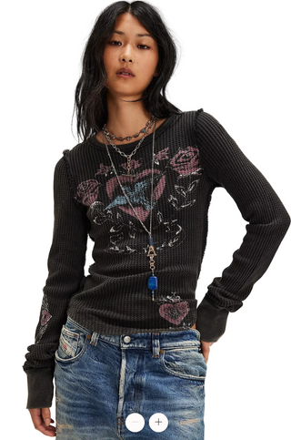 A woman modeling a black ribbed long-sleeve top with a graphic of hearts, roses, and a crown on the front, paired with blue jeans and layered necklaces, against a light gray background.