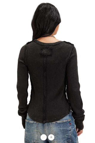 A woman shown from the back wearing a black ribbed long-sleeve top with distressed details and frayed edges, paired with blue jeans, against a plain light gray background.