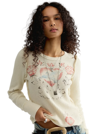 A woman with curly dark hair wearing a cream-colored waffle-knit sweater with a graphic of hearts, roses, and lips in pink, red, and blue, paired with blue jeans and a silver pendant necklace.