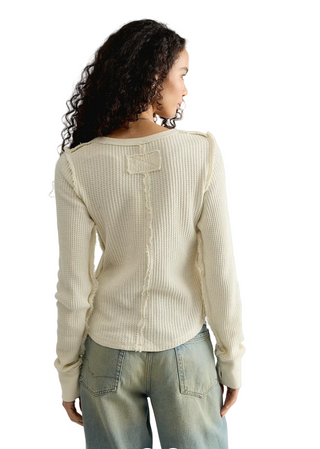 A woman shown from the back wearing a cream-colored ribbed knit sweater with a decorative lace-up detail down the center back, paired with light-wash jeans against a plain light gray studio background.
