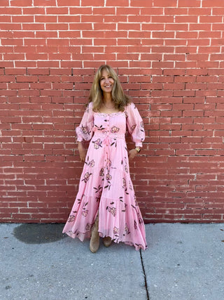 Woman in a pink floral dress standing against a red brick wall.