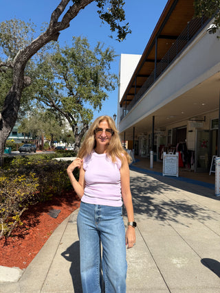 Woman in a pink top and blue jeans standing on a sidewalk with trees and a building in the background.