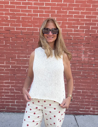 Woman wearing a white sleeveless top and polka dot pants standing in front of a red brick wall.
