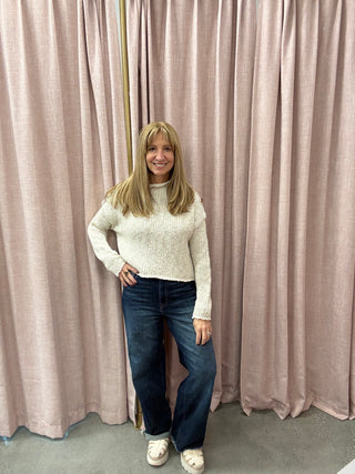 Woman wearing a white sweater and blue jeans standing in front of pink curtains.