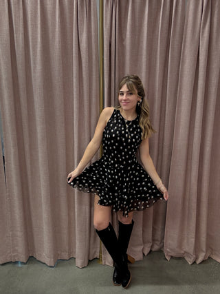 Woman in a black polka dot dress standing on a staircase with beige walls.