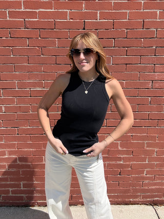 Woman wearing a black sleeveless top and white pants standing against a red brick wall.