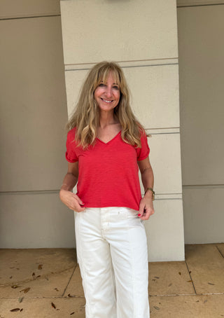 Woman wearing a red shirt and white pants standing against a beige wall.