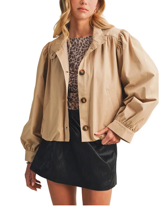 A woman models a beige cropped jacket with dramatic puffed sleeves and brown buttons, worn over a leopard-print top and a black mini skirt, against a plain beige studio background.