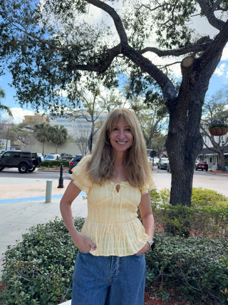 Woman in a yellow top and blue jeans standing outdoors with trees and parked cars in the background.