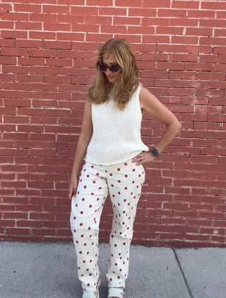 Woman wearing a white sleeveless top and polka dot pants standing against a red brick wall.