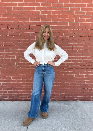 Woman in a white blouse and blue jeans standing against a red brick wall.