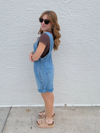 Person wearing a blue romper and sunglasses standing on a white wall.
