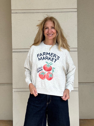 A woman wearing a light heather gray sweatshirt with a 'FARMER'S MARKET' graphic featuring red tomatoes, paired with dark blue jeans, standing in front of a beige stone wall.