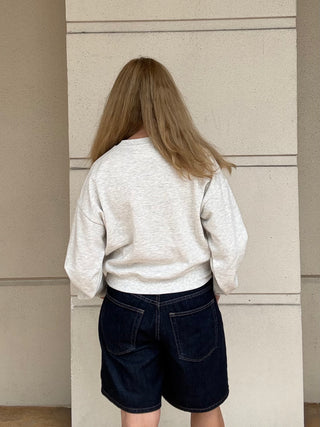 A person viewed from behind wearing a light heather gray long-sleeve crewneck sweatshirt and dark blue denim shorts, standing against a beige textured wall.
