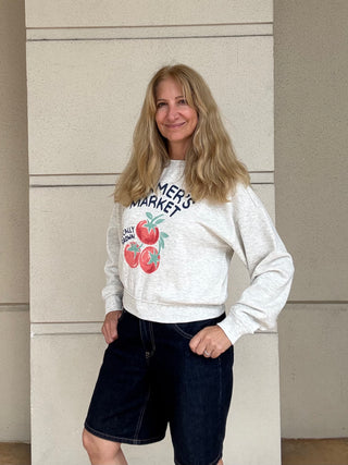 A woman with blonde hair stands against a beige wall wearing a light heather gray sweatshirt with a 'Summer's Market' graphic featuring red apples, paired with dark denim shorts.