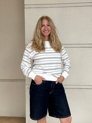 A smiling blonde woman stands against a beige wall wearing a white long-sleeve crewneck sweatshirt with thin dark horizontal stripes and dark denim shorts.