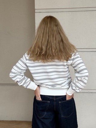 A woman viewed from behind wearing a white long-sleeve top with thin black horizontal stripes and dark blue jeans, standing against a neutral beige wall.