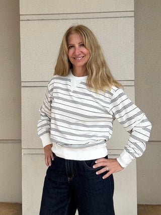 A woman with blonde hair wearing a white long-sleeve crewneck sweatshirt with thin black horizontal stripes and dark blue jeans, standing against a beige textured wall.