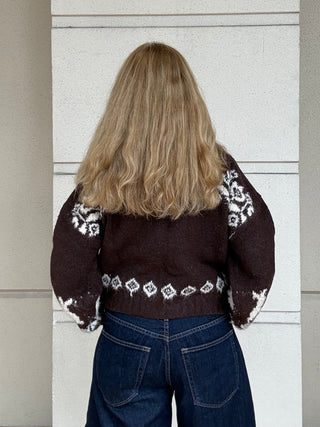 A person viewed from behind wearing a dark brown knit sweater with white Fair Isle patterns on the shoulders, back, and hem, paired with blue jeans, standing against a light gray paneled wall.