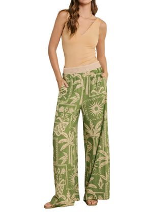 Woman wearing a beige top and green patterned pants against a white wall.