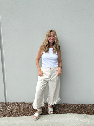 Woman wearing a white tank top and light-colored pants standing against a gray wall.
