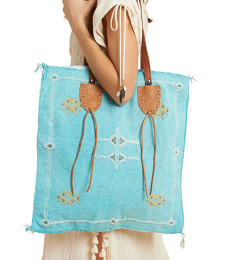 A turquoise blue woven tote bag with tan leather handles and fringe details, carried by a model against a solid beige background.
