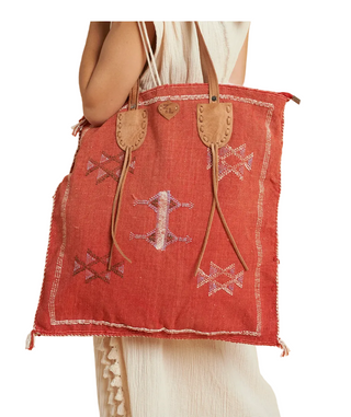 A terracotta red woven tote bag with tan leather handles and embroidered geometric patterns, held by a model wearing a cream dress against a solid beige background.